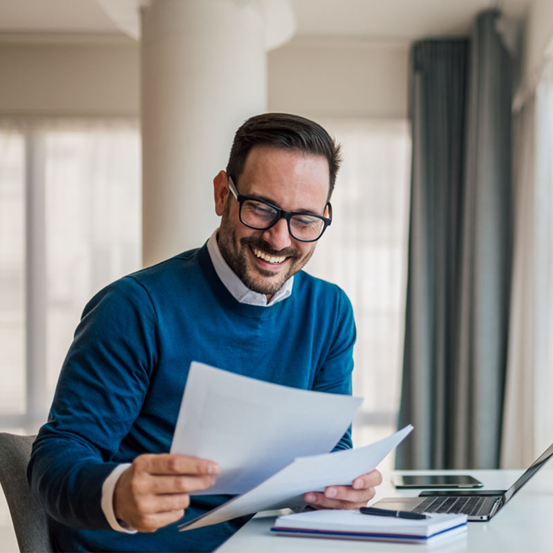 A man at a desk smiling while reading papers with a laptop and notebook.