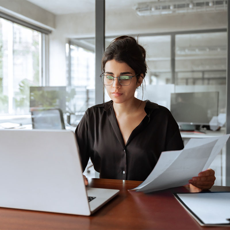 Woman in office working on laptop, holding documents.