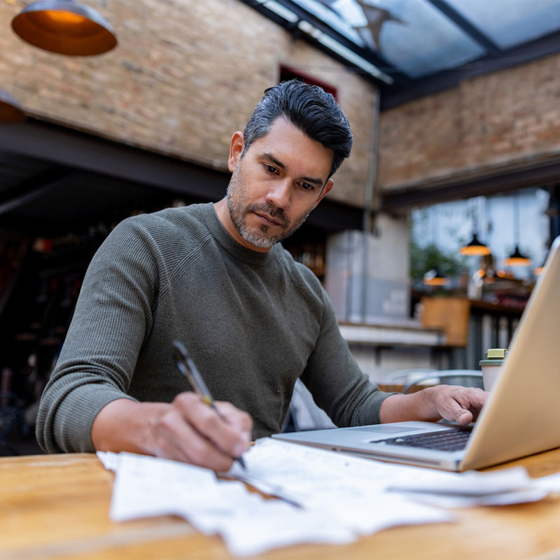 Man working on a laptop and writing notes at an outdoor café.