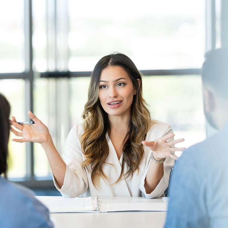 A woman in a business meeting gestures while speaking to two people across a table.