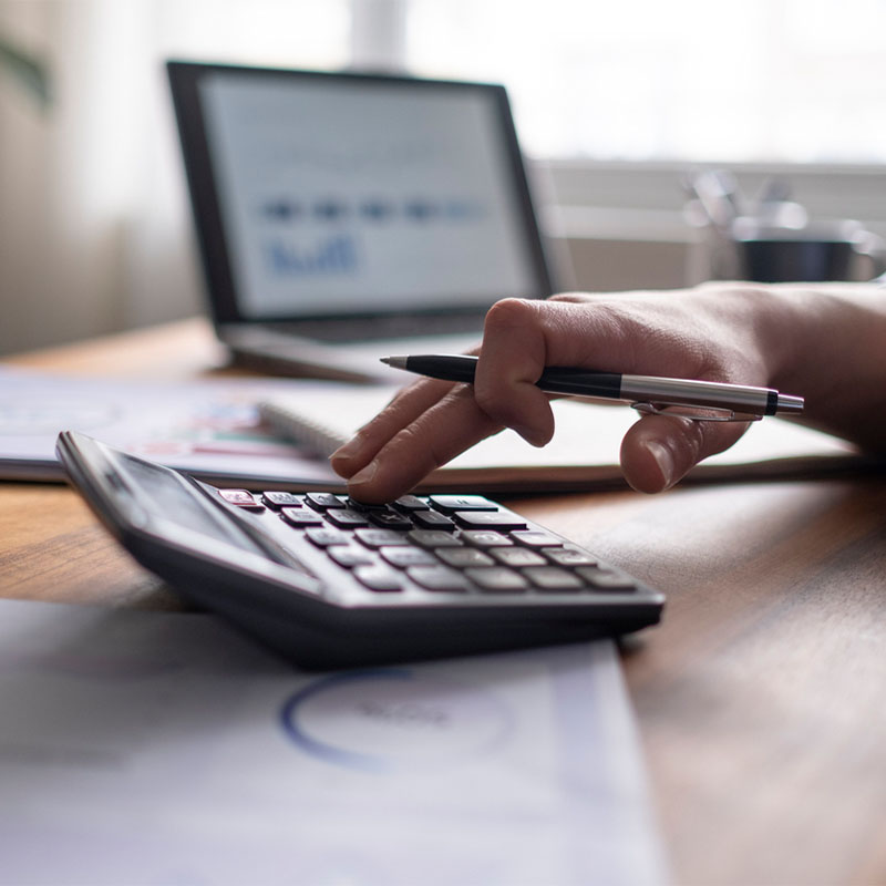 Hand using calculator with pen, laptop, and documents in background on office desk.