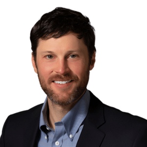 A man with dark hair and a beard wearing a blue shirt and blazer against a white background.