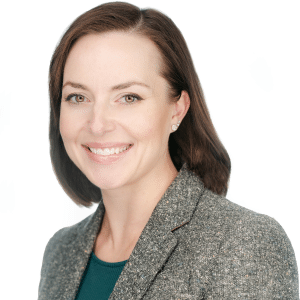 A woman smiling in a gray blazer and teal top against a white background.