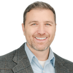 A smiling man in a gray blazer and blue shirt against a white background.