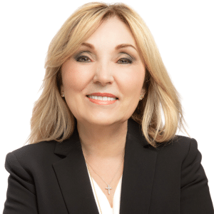 A smiling woman in a black blazer with blonde hair and a cross necklace.