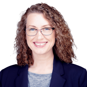 A smiling person with curly hair and glasses wearing a blazer against a white background.