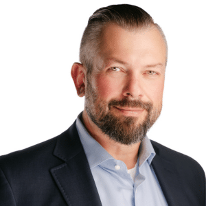 A man in a suit smiling with a neatly groomed beard and slicked-back hair.