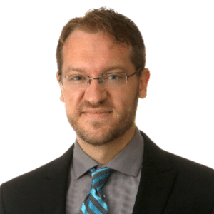 A person in a suit and glasses with a blue tie, standing against a white background.