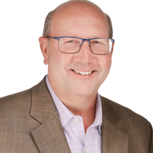 Man in glasses wearing a suit jacket and checkered shirt, smiling against a white background.