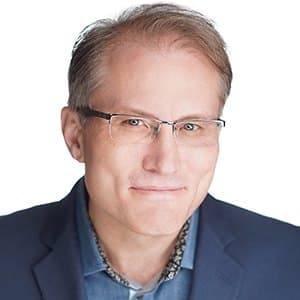 A man with glasses and a slight smile wearing a blue jacket against a white background.