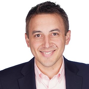 A smiling man in a suit and pink shirt with short dark hair against a white background.
