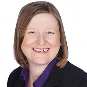 A woman smiling in a professional headshot, wearing a black blazer and purple shirt against a white background.