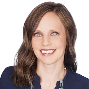 A woman with shoulder-length brown hair smiling, wearing a dark top and necklace against a white background.