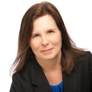 A woman in a black blazer with brown hair smiling against a white background.