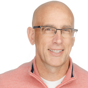 A man in glasses wearing a pink sweater smiles at the camera.