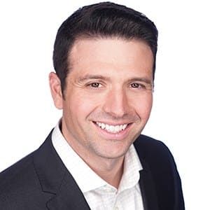 A smiling man in a suit with dark hair and a white shirt, against a white background.