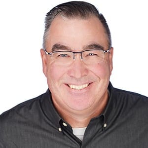A smiling man with glasses and gray shirt against a white background.