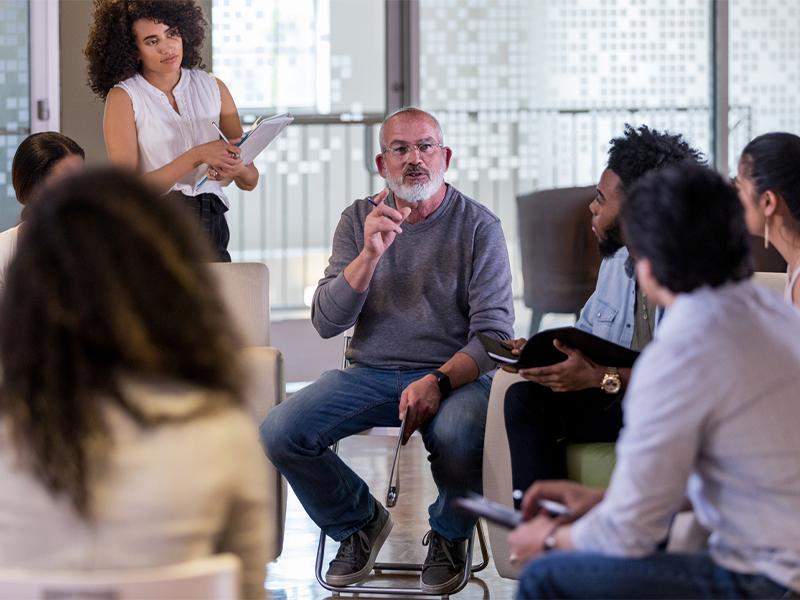 A group discussion with a man speaking and others listening attentively, one woman taking notes.